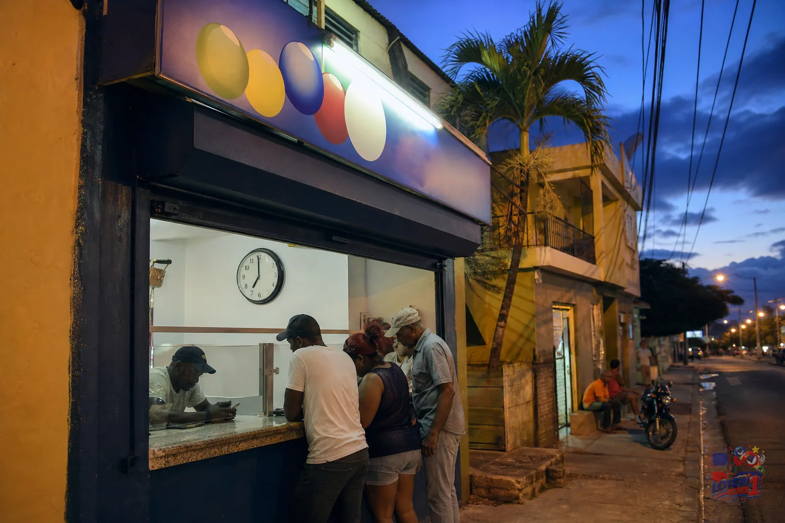 Banca de lotería dominicana al atardecer con un reloj que marca las siete de la noche, hora del sorteo nocturno de La Primera.